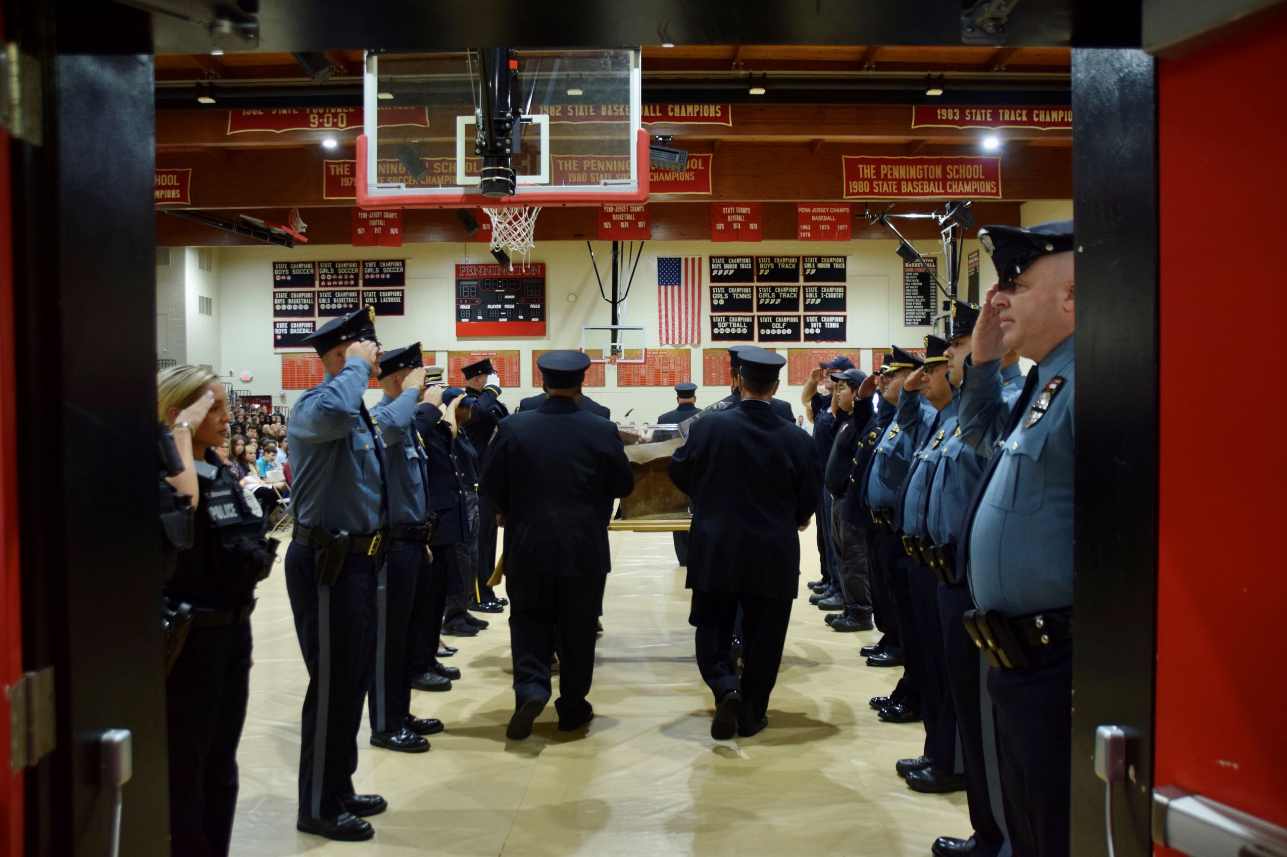 Photo of 4 firefighters carrying a piece of steel into a gym surrounded by saluting fire and police