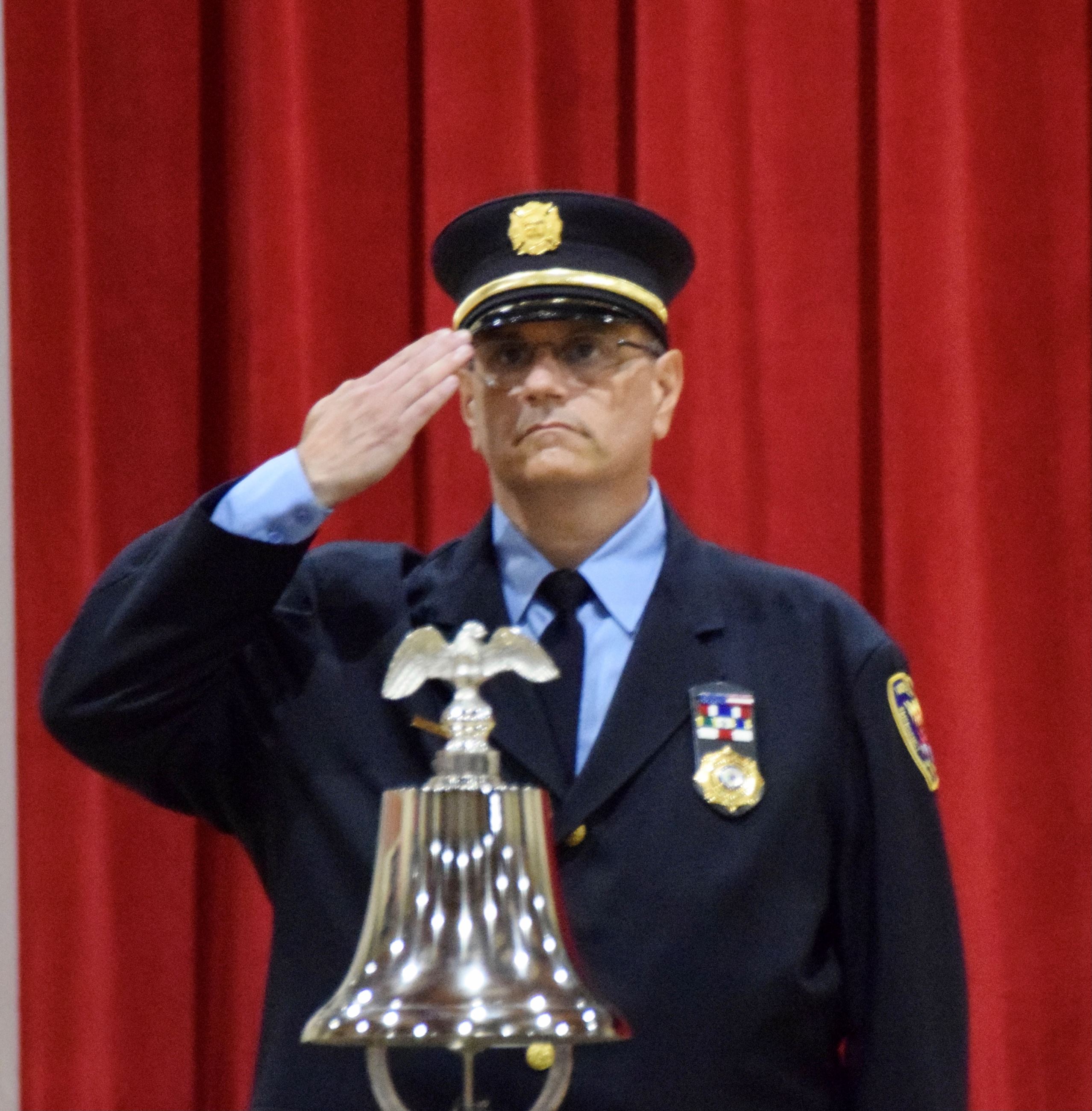 Firefighter in dress uniform saluting with bell in front. Bell has an eagle on top.